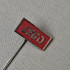 Puede incluir: Un pin rectangular con la palabra "LEGO" en letras plateadas sobre un fondo rojo. El pin tiene un vástago plateado y es probablemente un objeto de colección o un accesorio.