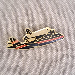 May include: A gold-colored pin shaped like a space shuttle, with black and red accents. The pin features the text "MASA" on the left wing. The shuttle design includes black lines and a red stripe along the bottom.