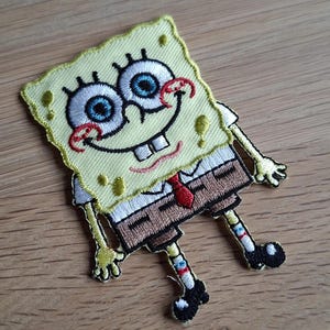 May include: Embroidered patch featuring the character SpongeBob SquarePants. The patch has a yellow sponge body, a white shirt, brown pants, and a red tie. The eyes are large and blue, and the mouth is smiling.