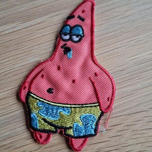 May include: Embroidered patch featuring Patrick Star from Spongebob Squarepants. The patch is a pink starfish shape with blue and yellow shorts. Patrick has a droopy expression with blue eyes and a blue drool.