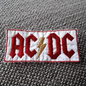 Peut inclure: &Eacute;cusson brod&eacute; avec le texte "AC/DC" en lettres gothiques rouges et un &eacute;clair dor&eacute; sur fond blanc. L'&eacute;cusson a une bordure rouge et est de forme rectangulaire.