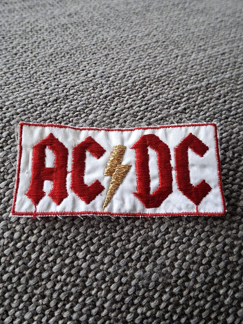 Peut inclure: &Eacute;cusson brod&eacute; avec le texte "AC/DC" en lettres gothiques rouges et un &eacute;clair dor&eacute; sur fond blanc. L'&eacute;cusson a une bordure rouge et est de forme rectangulaire.
