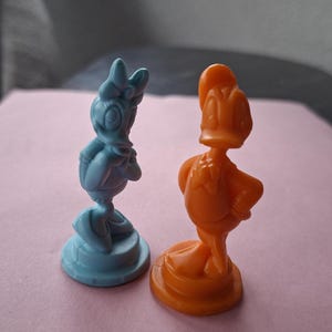 May include: Two small, plastic figurines of cartoon characters. One is blue and resembles Daisy Duck, the other is orange and resembles Donald Duck. Both figures are standing on round bases.