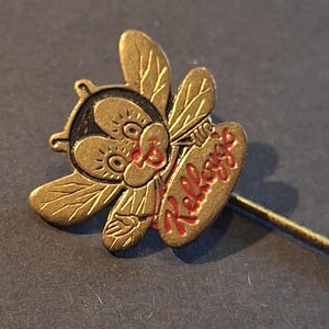 Op de afbeelding: Een vintage speld met een gestileerde bij met het woord "Kellogg's" in het rood. De speld heeft een goudkleurige afwerking en een lange, dunne speld. De bij heeft grote ogen en gedetailleerde vleugels.