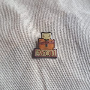 May include: A gold and orange Avon pin with the word "Sensual" in a pink banner and the word "Avon" in a gold banner.