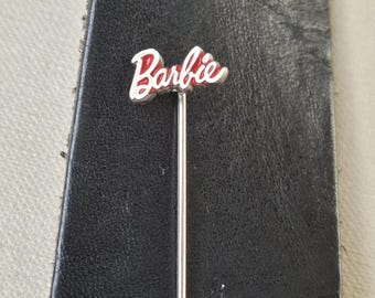 Op de afbeelding: Een zilverkleurige speld met het woord "Barbie" in rode letters met een witte omlijning. De speld staat tegen een zwarte achtergrond.