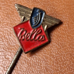 May include: A vintage Bella pin badge with a red diamond-shaped emblem and gold lettering. The pin features a blue and gold crest and gold wings. The pin is made of metal and has a long, thin pin backing.