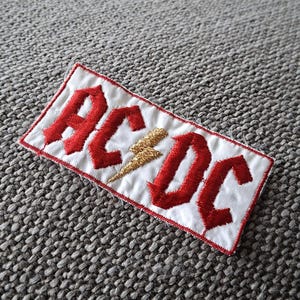 Puede incluir: Parche bordado con el nombre de la banda "AC/DC" en letras góticas rojas sobre fondo blanco. Un rayo dorado se encuentra entre las letras. El parche tiene un borde rojo, ideal para ropa o accesorios.