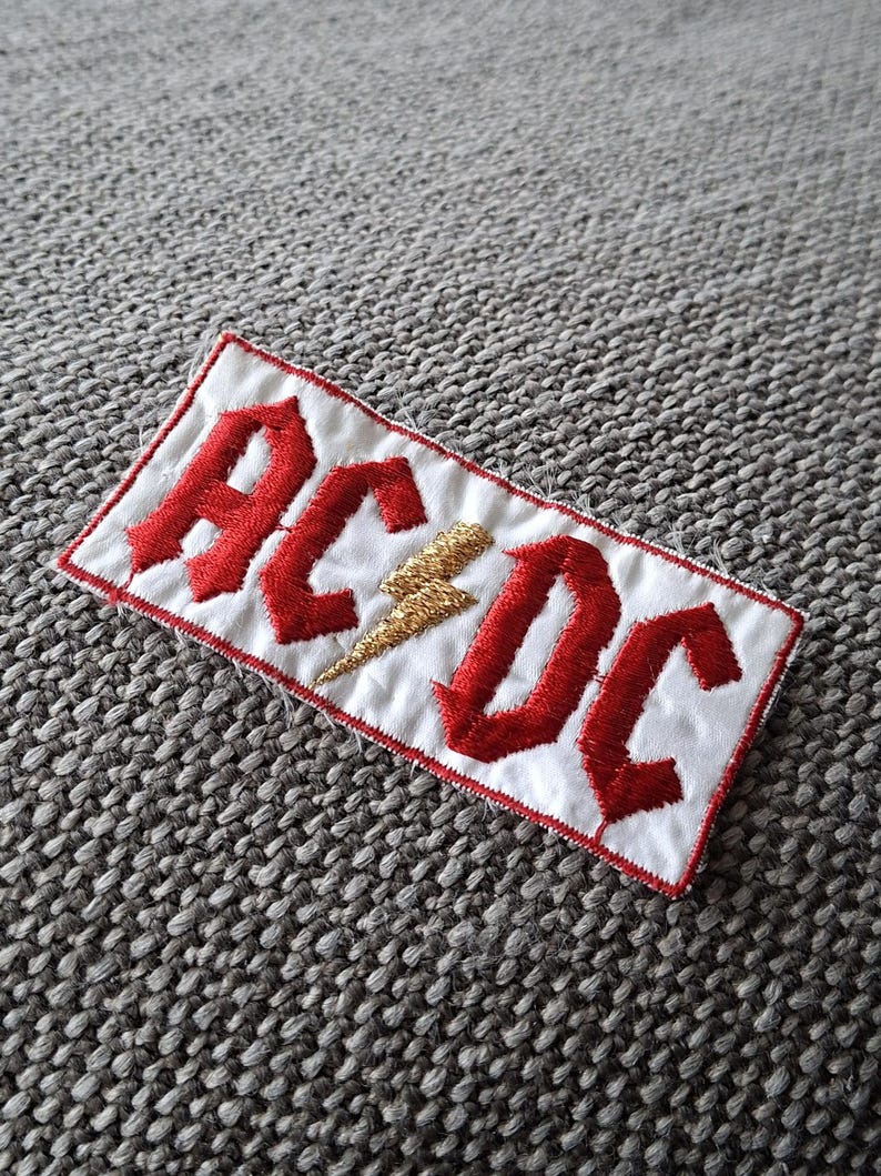 Peut inclure: &Eacute;cusson brod&eacute; avec le nom du groupe "AC/DC" en lettres gothiques rouges sur fond blanc. Un &eacute;clair dor&eacute; est plac&eacute; entre les lettres. Le patch a une bordure rouge, id&eacute;al pour les v&ecirc;tements ou accessoires.