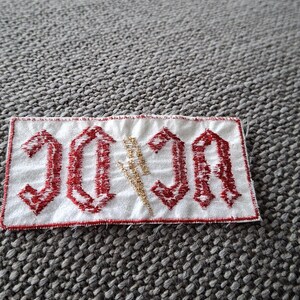 Peut inclure: Un &eacute;cusson rectangulaire blanc avec le texte "AC/DC" en rouge, dans une police gothique. Un &eacute;clair dor&eacute; est plac&eacute; entre les lettres. Le patch est bord&eacute; de rouge.
