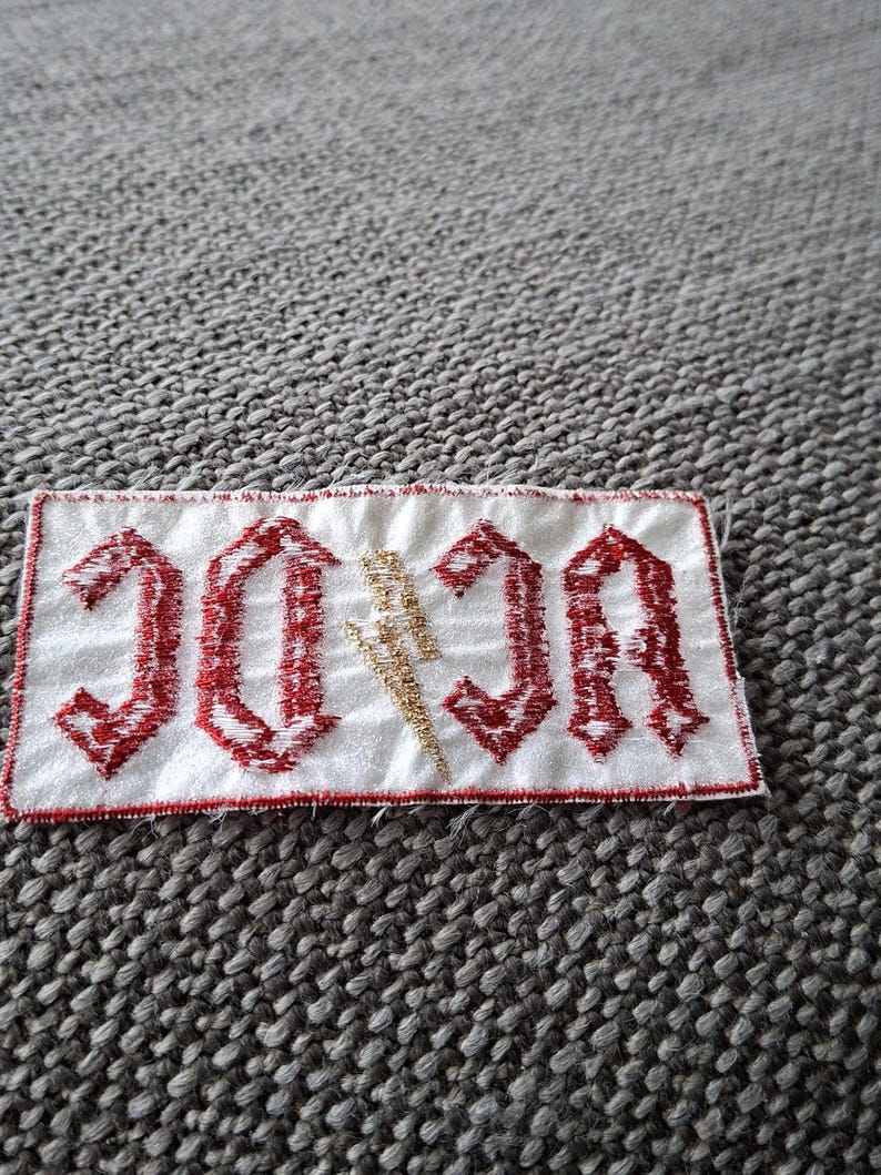 Peut inclure: Un &eacute;cusson rectangulaire blanc avec le texte "AC/DC" en rouge, dans une police gothique. Un &eacute;clair dor&eacute; est plac&eacute; entre les lettres. Le patch est bord&eacute; de rouge.
