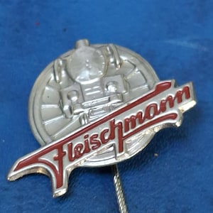 May include: A silver-colored Fleischmann pin with a red and white logo. The pin features a train wheel design and the brand name in a stylized font. The pin has a silver-colored pin backing.