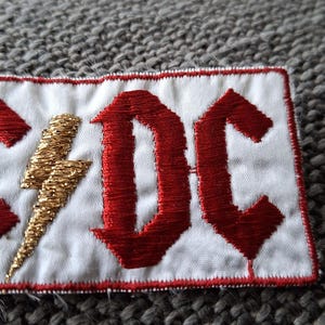 Peut inclure: &Eacute;cusson brod&eacute; avec les lettres "AC/DC" en rouge, de style gothique, et un &eacute;clair dor&eacute; sur fond blanc. Le patch a une bordure rouge, id&eacute;al pour les v&ecirc;tements ou accessoires.
