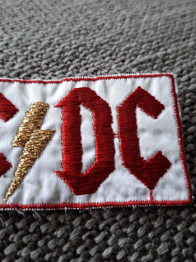 Peut inclure: &Eacute;cusson brod&eacute; avec les lettres "AC/DC" en rouge, de style gothique, et un &eacute;clair dor&eacute; sur fond blanc. Le patch a une bordure rouge, id&eacute;al pour les v&ecirc;tements ou accessoires.
