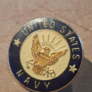 Puede incluir: Pin redondo con un emblema de águila dorada, rodeado por las palabras "UNITED STATES NAVY" en un borde azul oscuro. El pin tiene un centro dorado con el diseño del águila y rayos de sol, y dos estrellas doradas.