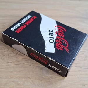 May include: A rectangular deck of playing cards in a black box featuring the Coca-Cola Zero logo, a white bottle silhouette, and the red text PLAYING POKER WITHOUT LOSING. The side of the box displays the Coca-Cola zero brand name in red and white.