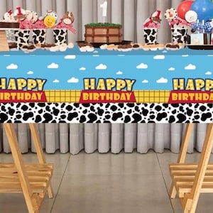 May include: A blue tablecloth with a cartoon design, featuring the text "HAPPY BIRTHDAY" and a black and white cow print. The table is set with cupcakes, a cake, and balloons, creating a festive atmosphere for a birthday celebration.