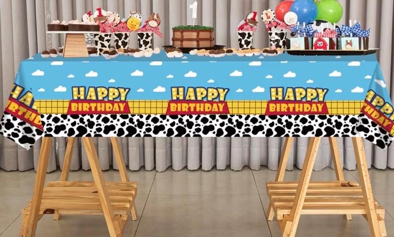 May include: A blue tablecloth with a cartoon design, featuring the text "HAPPY BIRTHDAY" and a black and white cow print. The table is set with cupcakes, a cake, and balloons, creating a festive atmosphere for a birthday celebration.
