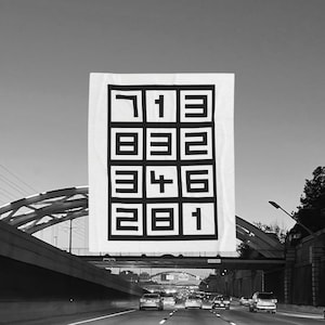Puede incluir: Una gran pancarta blanca con una cuadrícula negra de números y símbolos está suspendida sobre una autopista. Los números están dispuestos en una cuadrícula de 3x3. La pancarta cuelga de un puente, con coches debajo.