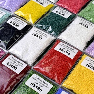 May include: Assortment of small, colorful seed beads in clear plastic bags. Each bag has a label with the brand name "Preciosa Ornela", "Made in Czech Republic", and the size "10/0". The bags are arranged in rows, with the labels facing the viewer. The beads are in various colors, including red, green, white, black, yellow, and blue.