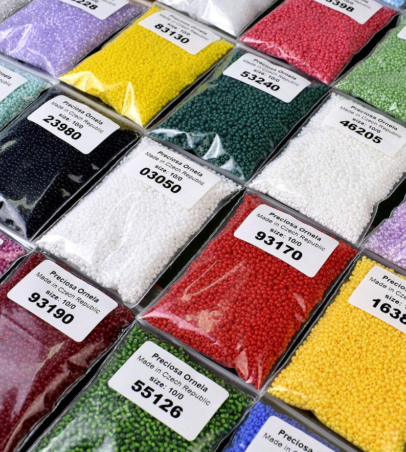 May include: Assortment of small, colorful seed beads in clear plastic bags. Each bag has a label with the brand name "Preciosa Ornela", "Made in Czech Republic", and the size "10/0". The bags are arranged in rows, with the labels facing the viewer. The beads are in various colors, including red, green, white, black, yellow, and blue.