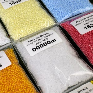 May include: Close-up of six bags of small, round beads. The bags are labeled with the brand name "Preciosa Ornela" and the size of the beads. The beads are in various colors, including yellow, white, light blue, and pink.