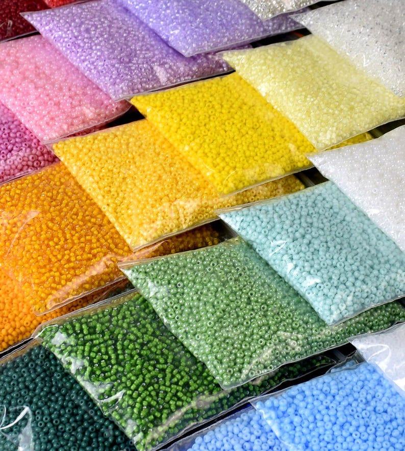 May include: Assortment of seed beads in various colors, including light pink, lavender, yellow, orange, green, blue, and white. The beads are packaged in clear plastic bags.