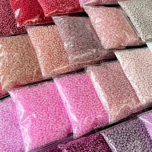 Op de afbeelding: Assortiment van kleine, doorzichtige plastic zakjes gevuld met verschillende tinten roze en witte kralen. De kralen zijn in rijen gerangschikt, wat een visueel aantrekkelijk display creëert.