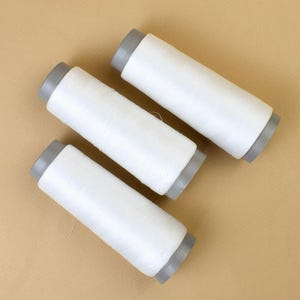 Può includere: Tre rocchetti di filo bianco con tappi grigi sono disposti in diagonale. Il filo è ben avvolto su rocchetti cilindrici. L'immagine è ben illuminata, mostrando la trama del filo. Sfondo beige.