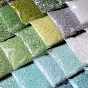 May include: Assortment of small, round glass beads in various shades of green, blue, and white, packaged in clear plastic bags. The beads are arranged in a grid pattern, creating a visually appealing display.