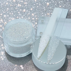 May include: A clear jar filled with iridescent white glitter, along with two clear nail tips covered in the same glitter. The glitter is on a silver background.
