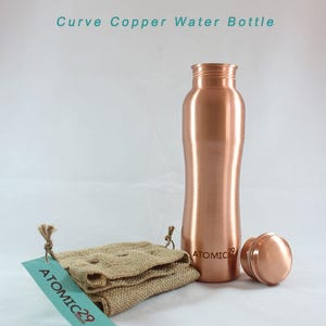K&ouml;nnte beinhalten: Eine Wasserflasche aus Kupfer mit einem geschwungenen Design. Die Flasche hat einen Deckel und steht auf einer wei&szlig;en Oberfl&auml;che. Die Flasche ist mit "ATOMIC 29" beschriftet.
