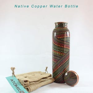 K&ouml;nnte beinhalten: Eine Kupfer-Wasserflasche mit einem schwarzen und braunen Muster. Die Flasche hat einen Kupferdeckel und wird mit einem Jutesack geliefert. Der Sack hat ein Etikett mit der Aufschrift "ATOMIC29".