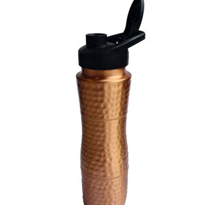 K&ouml;nnte beinhalten: Eine geh&auml;mmerte Kupfer-Wasserflasche mit einem schwarzen Pop-Top-Deckel. Die Flasche hat eine strukturierte, geh&auml;mmerte Oberfl&auml;che.