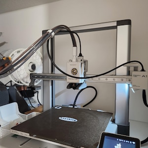 Puede incluir: Una impresora 3D plateada y negra con el texto "Bambu Lab A1" en el lateral. La impresora tiene una cama de impresión negra con la palabra "Anything" encima. Una pequeña pantalla muestra la configuración de la impresora.