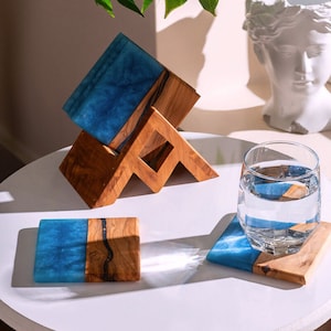 Puede incluir: Un juego de posavasos rectangulares hechos de madera natural y resina azul, con un soporte de madera para almacenamiento. Un posavasos está colocado plano, mientras que otro sostiene un vaso transparente lleno de agua. Se aprecia la veta.