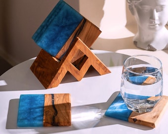 May include: A set of rectangular coasters made from natural wood and blue resin, featuring a wooden storage stand. One coaster lies flat, while another supports a clear glass filled with water. The wood grain is visible alongside the blue resin.