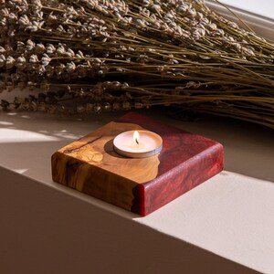 May include: A square wooden candle holder with a red and natural wood finish. A small white candle is burning in the center of the holder.