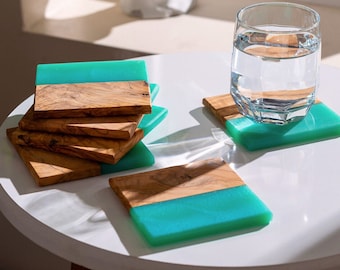 May include: A set of square coasters featuring a combination of natural brown wood grain and translucent bright turquoise resin. One coaster is placed separately with a clear glass of water on top, while a stack of four coasters sits nearby.