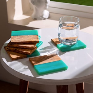 Può includere: Un set di sottobicchieri fatti a mano con un design moderno. Ogni sottobicchiere presenta una forma rettangolare con una sezione in legno naturale e una sezione in resina turchese vibrante. Un bicchiere d'acqua è appoggiato su uno dei sottobicchieri.