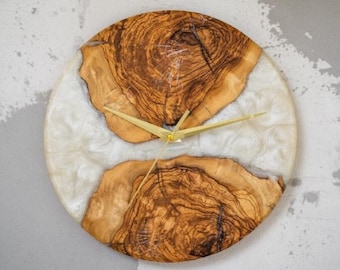 Puede incluir: Un reloj de pared circular que presenta dos piezas de madera natural con patrones de veta prominentes separados por una banda de resina blanca translúcida. El reloj tiene manecillas de metal dorado fino centradas en la esfera.