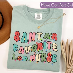 May include: A light green t-shirt with the text "SANTA'S FAVORITE L&D NURSE" in colorful, retro-style lettering. The design includes a string of Christmas lights. The shirt is a Comfort Colors brand.