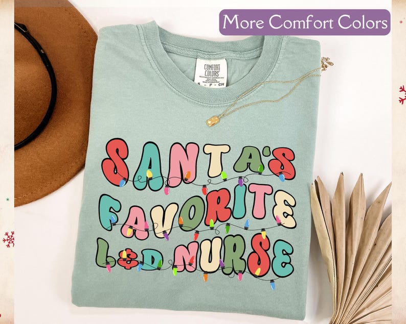 May include: A light green t-shirt with the text "SANTA'S FAVORITE L&D NURSE" in colorful, retro-style lettering. The design includes a string of Christmas lights. The shirt is a Comfort Colors brand.