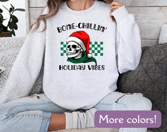 May include: A light gray sweatshirt with a graphic of a skull wearing a Santa hat and green scarf. The text "BONE-CHILLIN' HOLIDAY VIBES" is above and below the skull. The image also includes a green and white checkered pattern.