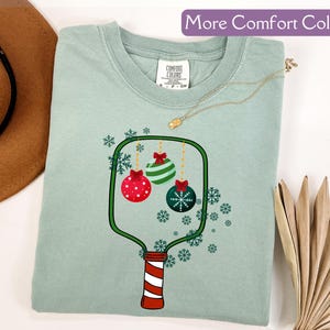 Puede incluir: Una camiseta verde claro con un dise&ntilde;o de adorno navide&ntilde;o. El gr&aacute;fico presenta tres adornos en rojo, verde y azul, con copos de nieve. El texto "More Comfort Colors" est&aacute; en la parte superior.