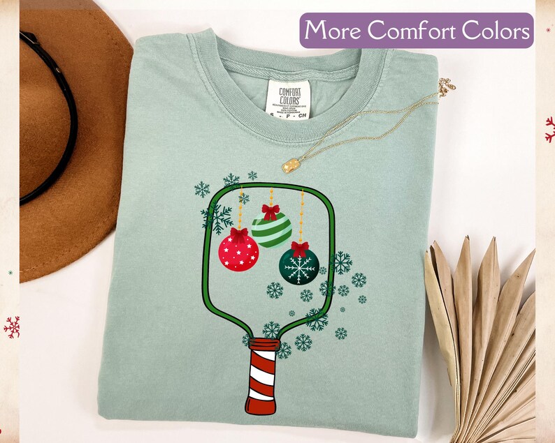 Puede incluir: Una camiseta verde claro con un dise&ntilde;o de adorno navide&ntilde;o. El gr&aacute;fico presenta tres adornos en rojo, verde y azul, con copos de nieve. El texto "More Comfort Colors" est&aacute; en la parte superior.