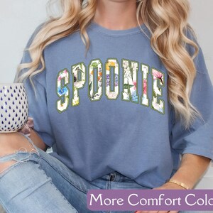 K&ouml;nnte beinhalten: Ein blaues T-Shirt mit dem Wort "SPOONIE" in wei&szlig;en Buchstaben mit einem Blumenmuster. Das Shirt wird von einer Person mit langen blonden Haaren getragen.