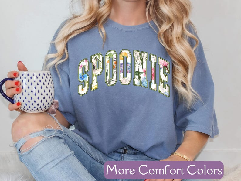 K&ouml;nnte beinhalten: Ein blaues T-Shirt mit dem Wort "SPOONIE" in wei&szlig;en Buchstaben mit einem Blumenmuster. Das Shirt wird von einer Person mit langen blonden Haaren getragen.