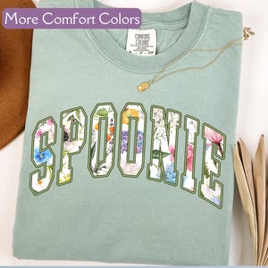 K&ouml;nnte beinhalten: Ein hellgr&uuml;nes T-Shirt mit dem Wort "SPOONIE" in wei&szlig;en, gr&uuml;n umrandeten Buchstaben. Die Buchstaben sind mit einem Blumenmuster gef&uuml;llt. Das Shirt ist aus einem weichen, bequemen Stoff.