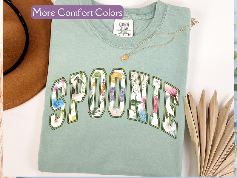K&ouml;nnte beinhalten: Ein hellgr&uuml;nes T-Shirt mit dem Wort "SPOONIE" in wei&szlig;en, gr&uuml;n umrandeten Buchstaben. Die Buchstaben sind mit einem Blumenmuster gef&uuml;llt. Das Shirt ist aus einem weichen, bequemen Stoff.